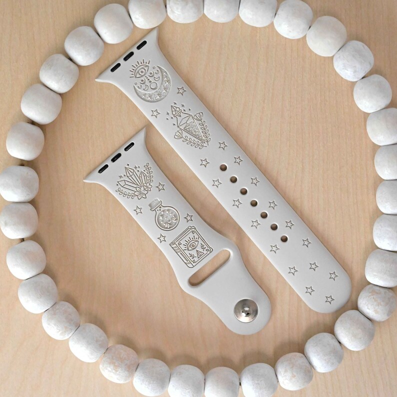 May include: Two gray Apple Watch bands with white engraved designs. One band features a crescent moon, a star, a crystal, and a potion bottle. The other band features a star pattern.