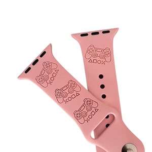 Può includere: Cinturino rosa in silicone per Apple Watch con un motivo ripetuto di un controller per videogiochi e i simboli X, O, quadrato e triangolo.