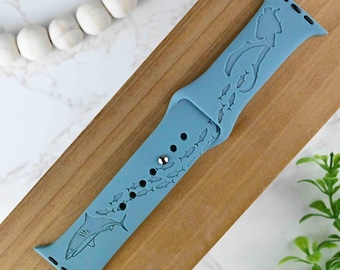 May include: A light blue silicone Apple Watch band with a shark and school of fish design. The band is made for a 40mm or 44mm Apple Watch.