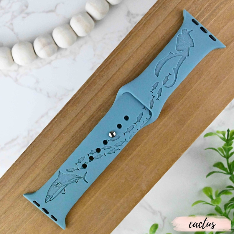 Op de afbeelding: Een lichtblauw siliconen Apple Watch-bandje met een haai- en vissenmotief. Het bandje is geschikt voor een Apple Watch van 40 mm of 44 mm.