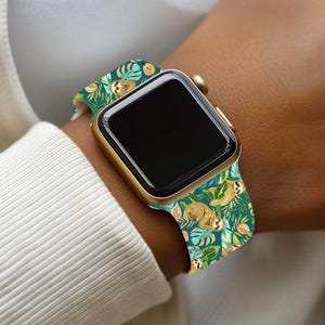 Puede incluir: Un Apple Watch dorado con una correa turquesa con un patrón de perezosos y hojas tropicales. La correa del reloj tiene un diseño repetido de perezosos, hojas y flores en tonos marrones, verdes y naranjas. La esfera del reloj es negra.
