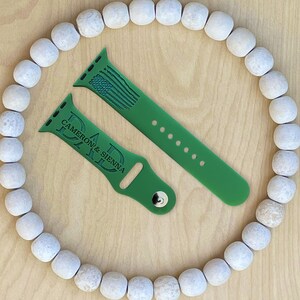 Puede incluir: Dos correas de reloj de silicona verde con un mensaje personalizado. Una correa tiene el texto "DAD" con los nombres "CAMERON & SIENNA" debajo. La otra correa tiene un peque&ntilde;o dise&ntilde;o de bandera estadounidense.