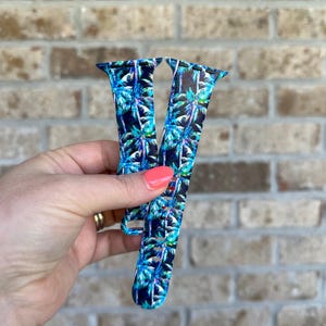 May include: A blue and black watch strap with a tropical palm tree design. The strap is held up against a brick wall. The watch strap is made of a flexible material and has a buckle fastening.