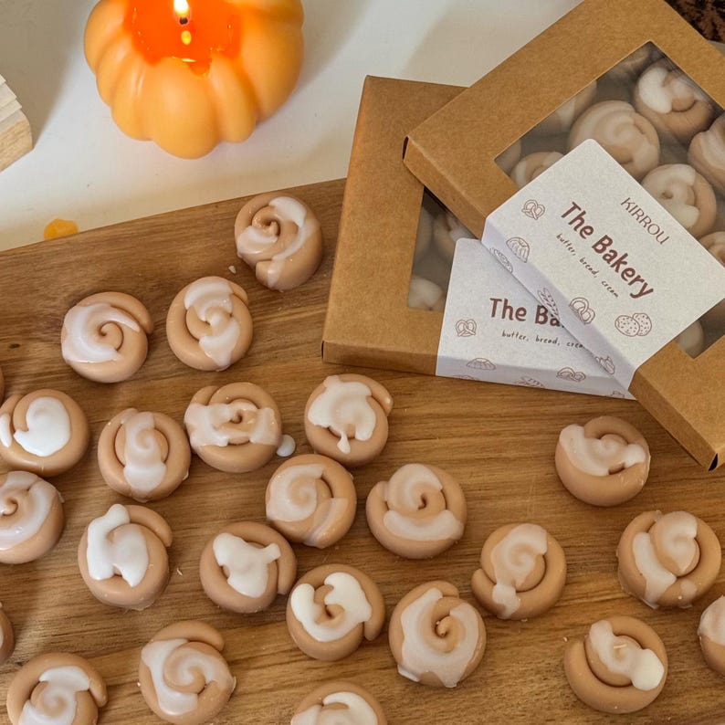 May include: A wooden cutting board displays cinnamon roll-shaped wax melts with white icing. Two boxes with clear lids are visible, one open, filled with similar melts. The text "The Bakery" is on the box. An orange pumpkin-shaped candle burns in the background.