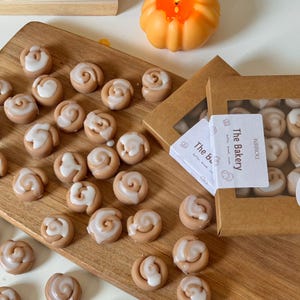 May include: A wooden cutting board displays several miniature cinnamon roll-shaped wax melts, each topped with white icing. The melts are arranged around two small boxes with the text "The Bakery" on the label. A small orange pumpkin decoration is in the background.