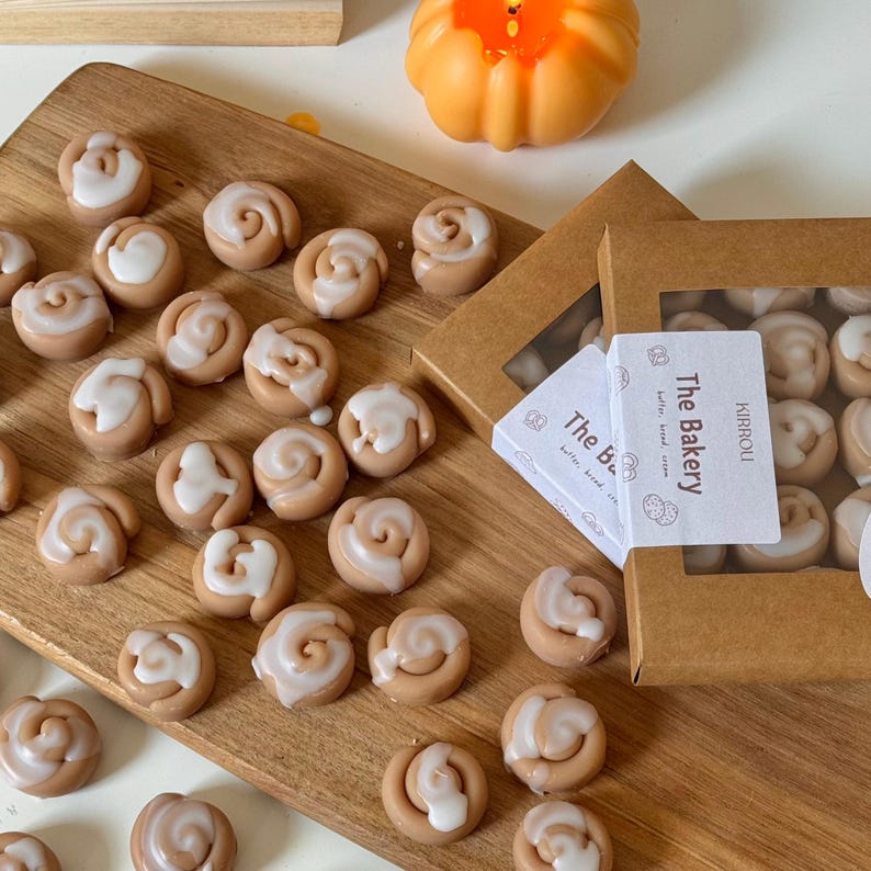 May include: A wooden cutting board displays several miniature cinnamon roll-shaped wax melts, each topped with white icing. The melts are arranged around two small boxes with the text "The Bakery" on the label. A small orange pumpkin decoration is in the background.