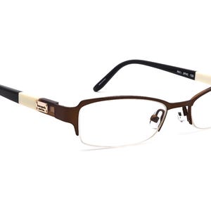 Könnte beinhalten: Braune und goldfarbene Brille mit halb rahmenlosem Design. Die Fassungen haben eine rechteckige Form mit einem dünnen Metallsteg und Bügeln. Die Bügel sind schwarz mit einem cremefarbenen Akzent in der Nähe der Scharniere. Die Gläser sind klar.