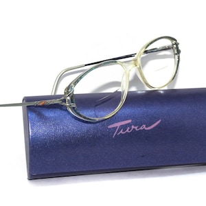 Puede incluir: Un par de gafas transparentes de forma ovalada con montura plateada y estuche azul y plateado. El estuche tiene la palabra "Tura" impresa en rosa en la parte delantera.