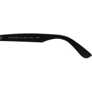 May include: Close-up of the black plastic arm of a pair of Wayfarer sunglasses. The arm is inscribed with the text "WAYFARER&reg; Hand Made in Italy K CE".