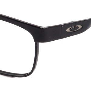 K&ouml;nnte beinhalten: Schwarze rechteckige Brille mit einem silbernen Oakley-Logo an der Seite.