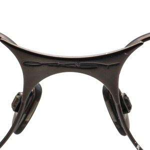 May include: Close-up of a pair of brown-toned sunglasses. The frame features a curved design with the brand name "OAKLEY" embossed on the bridge. The nose pads are visible, along with the temples extending outwards.