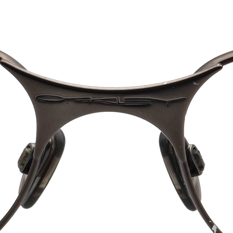 May include: Close-up of a pair of brown-toned sunglasses. The frame features a curved design with the brand name "OAKLEY" embossed on the bridge. The nose pads are visible, along with the temples extending outwards.