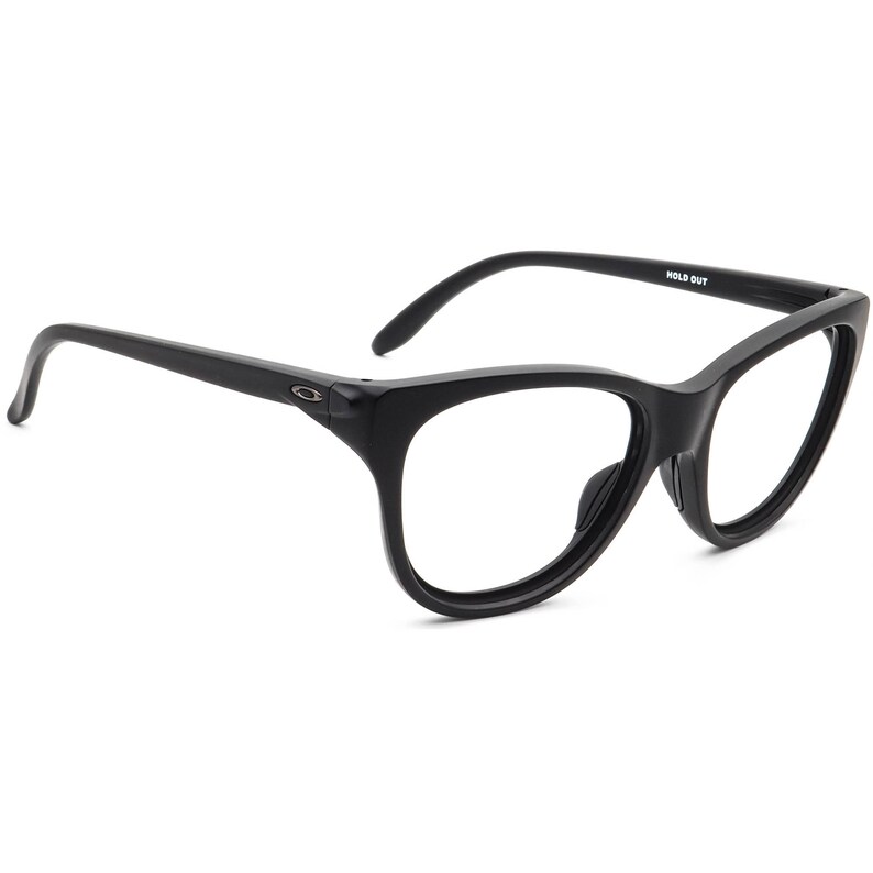 May include: Black cat-eye eyeglasses with clear lenses. The frames are thick and have a rounded shape. The text "HOLD OUT" is printed on the top of the frame. The Oakley logo is visible on the side of the frame.