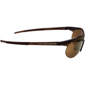 May include: Brown Oakley sunglasses with a textured frame and brown lenses. The brand name "Oakley" is printed in gold on the frame.