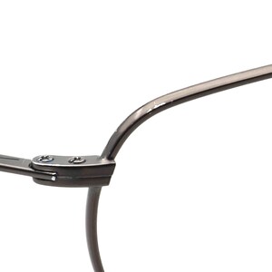 May include: Close-up of a section of eyeglasses. The frame is a dark gray metal with a curved design. The hinge is visible, with small screws securing the frame components. The background is plain white.