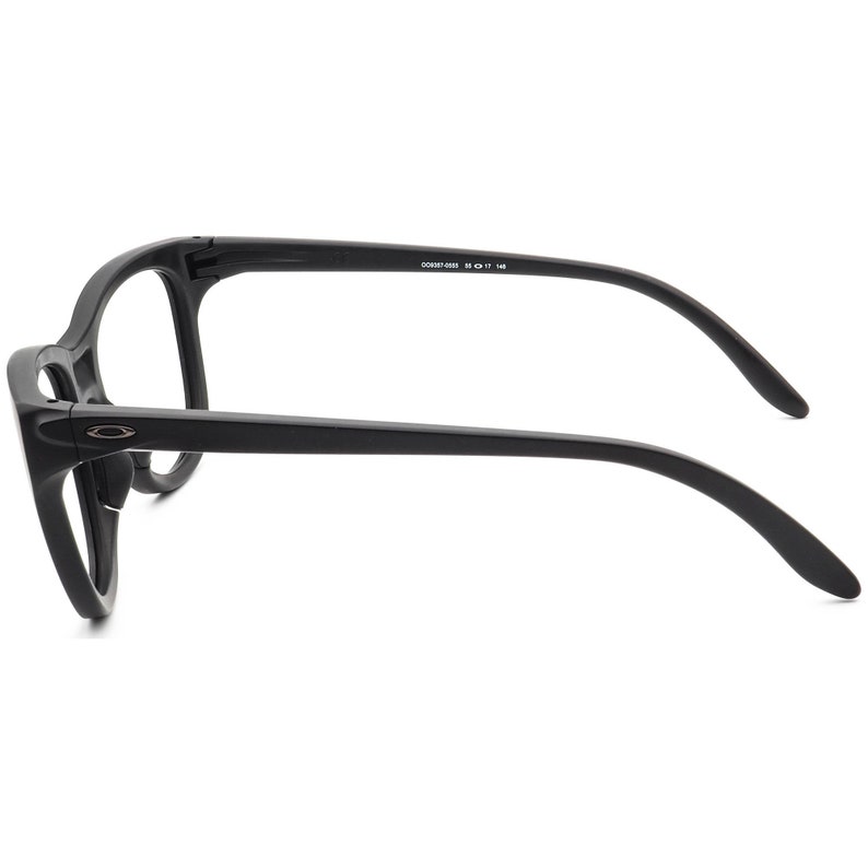 May include: Black eyeglasses with a matte finish. The glasses have a thick frame and curved temples. The Oakley logo is visible on the side of the frame. The text "OO9287-0855 55 17 145" is printed on the temple.