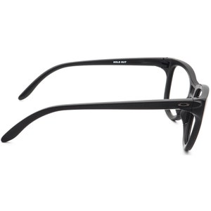May include: Black eyeglasses with a thick frame. The glasses have a rectangular shape and curved temples. The text "HOLD OUT" is printed on the left temple. The Oakley logo is visible on the right side of the frame.