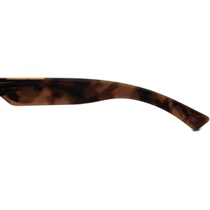 May include: Close-up of the tortoiseshell brown plastic temple arm of a pair of sunglasses. The arm is curved and has a small pink label that reads "USA Assembled".