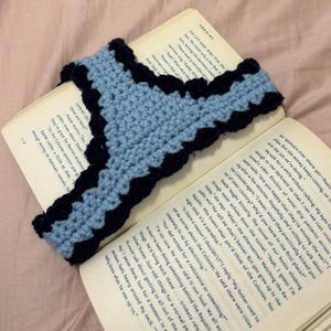 Pode incluir: Um marcador de crochê feito à mão em azul claro com uma borda ondulada azul escuro, repousando nas páginas de um livro aberto. O design do marcador é em forma de V estilizado.