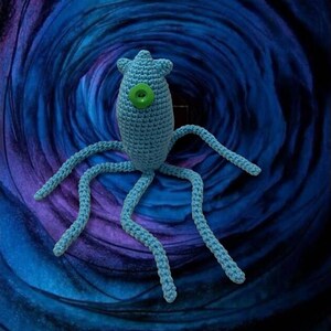 May include: A light blue crocheted creature with multiple tentacles and a green button eye. The handmade plush toy has a star-shaped head and is set against a swirling blue and purple background.