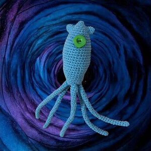 May include: A light blue crocheted sea creature with multiple tentacles and a large green button eye. The handmade plush toy is set against a swirling blue and purple background, creating a whimsical effect.