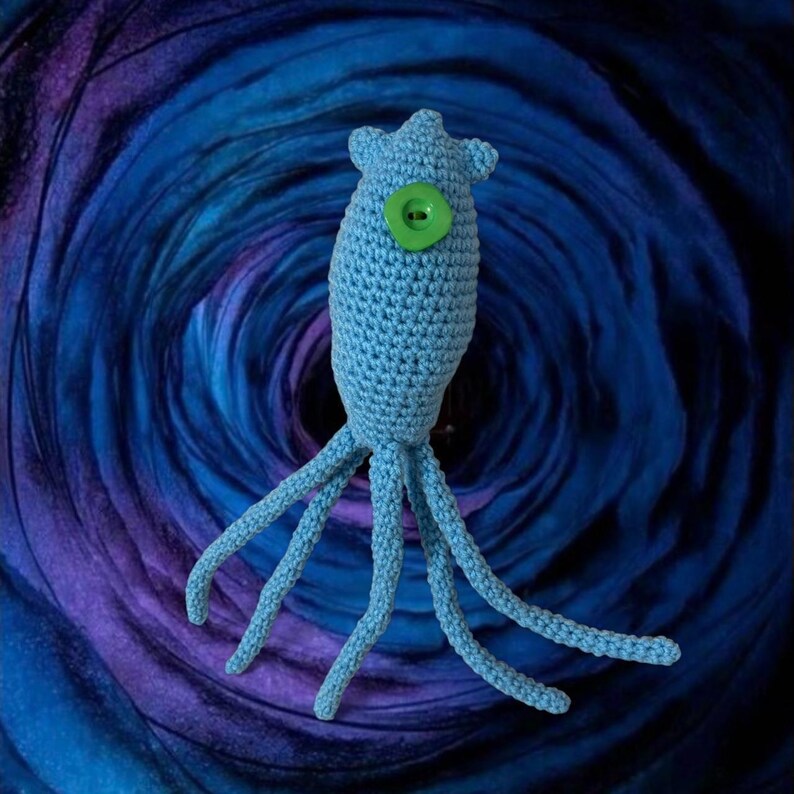 May include: A light blue crocheted sea creature with multiple tentacles and a large green button eye. The handmade plush toy is set against a swirling blue and purple background, creating a whimsical effect.