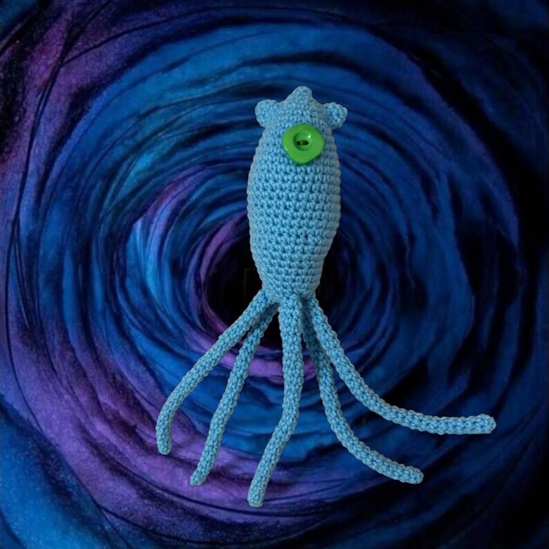 May include: A light blue crocheted creature with a green button eye and multiple tentacles. The handmade plush toy has a rounded body and a crown-like top. The background features a swirling pattern of blue and purple hues.