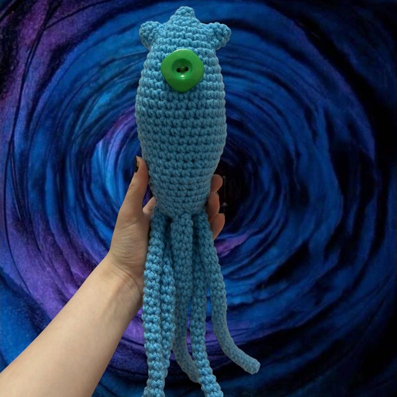 May include: A light blue crocheted squid plush toy with a large green button eye. The squid has multiple tentacles and is held against a swirling blue background. The handmade toy is soft and cuddly.