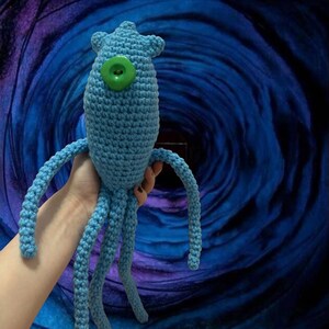 May include: A light blue crocheted creature with a large green button eye and multiple tentacles. The handmade plush toy has a textured surface and is set against a swirling blue and purple background.