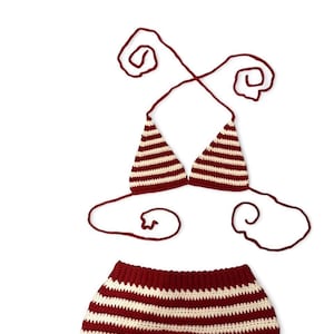 Puede incluir: Un conjunto de dos piezas de ganchillo compuesto por un top de bikini triangular y pantalones cortos a juego. Ambas piezas presentan rayas horizontales en rojo oscuro y blanco roto. El top tiene tiras finas y largas, y el pantalón una cintura ancha.