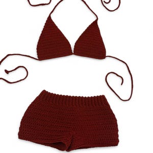 May include: A dark maroon two-piece crochet set consisting of a triangle bikini top with long adjustable tie strings and a pair of matching high-waisted shorts. The garment features a consistent textured stitch pattern throughout the entire design.