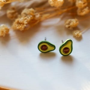 Può includere: Due orecchini a bottone a forma di avocado. Ogni orecchino presenta una buccia verde, un interno giallo e un seme marrone. Gli orecchini sono piccoli e poggiano su una superficie bianca. Un divertente accessorio a tema cibo.