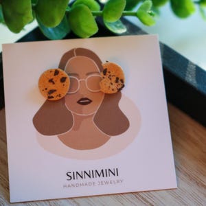 Könnte beinhalten: Handgefertigte Ohrringe in Keksform mit Schokoladenstückchen, präsentiert auf einer Karte mit einer Frauenillustration und dem Markennamen "SINNIMINI HANDMADE JEWELRY". Ein verspieltes und einzigartiges Accessoire.