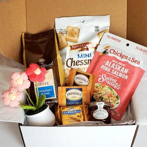 May include: A white gift box filled with a variety of food items, including coffee, tea, crackers, salmon, and honey. The box is decorated with pink flowers.