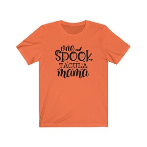 K&ouml;nnte beinhalten: Orangefarbenes T-Shirt mit dem schwarzen Schriftzug &bdquo;one spooktacular mama&ldquo;. Das Wort &bdquo;spook&ldquo; ist mit einer Fledermaus und einem Spinnennetz verziert. Das T-Shirt hat einen Rundhalsausschnitt und kurze &Auml;rmel.