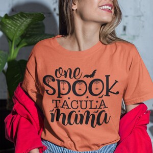 K&ouml;nnte beinhalten: Orangefarbenes T-Shirt mit dem schwarzen Text "one spooktacular mama", verziert mit Flederm&auml;usen und Spinnweben. Das Shirt wird mit blau-wei&szlig; gestreiften Jeans und einer roten Jacke getragen.