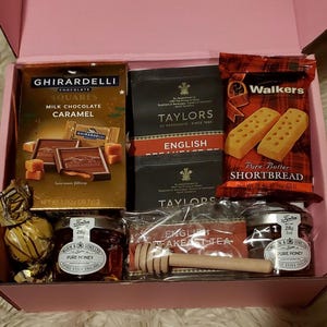 May include: A pink gift box filled with various treats, including a box of Ghirardelli milk chocolate caramel squares, a box of Taylors of Harrogate English Breakfast Tea, a package of Walkers Pure Butter Shortbread, two jars of honey, and a wooden honey dipper.