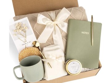 May include: A gift box filled with a beige blanket tied with a cream ribbon, a sage green mug, a notebook with "NOTES" printed on it, a gold pen, a small candle, a card, a gold spoon, a woven coaster, and a small white towel.