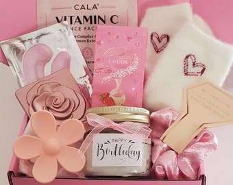 Pode incluir: Uma caixa de presente rosa e branca cheia de vários itens, incluindo um pote de sais de banho com um rótulo de "Feliz aniversário", um saboneteira em forma de rosa, uma flor rosa, uma máscara para os olhos, um pacote rosa e branco de chocolates Lindor, um chinelo branco felpudo com corações vermelhos bordados e uma escova de cabelo de madeira com o texto "Mantenha a calma e escove o cabelo para trás".