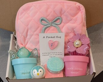 May include: A gift box filled with pink items. Includes a quilted cosmetic bag with a bow, a card that says "A Pocket Hug", a flower-shaped hair clip, and small containers of bath products. The box contains a bath bomb and a penguin-shaped item.
