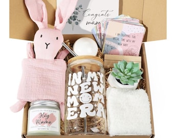 May include: A gift box filled with items for a new parent. The box contains a pink bunny toy, a glass jar with a straw and the text "In My New Mom Era", a candle with the text "Hey Mama", a small succulent plant in a white pot, a white bath bomb, and a card that says "Congrats Mam".