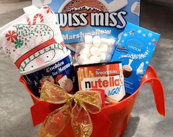 May include: A red plastic bucket filled with a Christmas-themed gift basket. The basket includes a mug that says "Cookies for Santa", a red and white striped kitchen towel, a box of Swiss Miss Marshmallow hot chocolate, a bag of marshmallows, a box of Nutella and breadsticks, and a bar of Lindor chocolate.