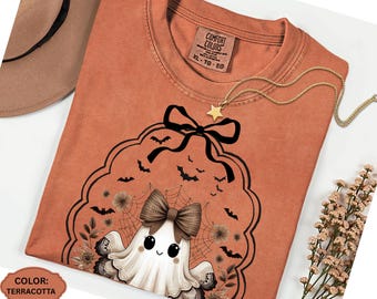May include: Terracotta-colored t-shirt featuring a Halloween-themed graphic. The design includes a cute ghost with a bow, bats, and floral elements. A gold star pendant necklace is draped on the shirt. The shirt has a Comfort Colors tag.