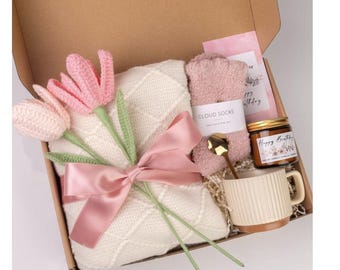 May include: A gift box filled with items for a cozy night in. It includes a white knitted blanket, pink cloud socks, a candle that says "Happy Birthday", and a mug. The box is decorated with a pink ribbon and three pink crocheted tulips.