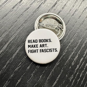 May include: Two white, round badges with the text "READ BOOKS. MAKE ART. FIGHT FASCISTS." in black. One badge is face up, and the other is face down, showing the pin back. The badges are on a dark wood surface.