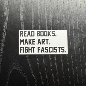 May include: A white sticker with black text stating "READ BOOKS. MAKE ART. FIGHT FASCISTS." The sticker is placed on a dark, textured surface, creating a contrast between the text and background.