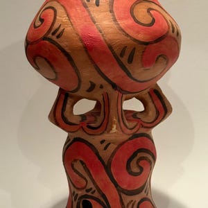 May include: A hand-painted ceramic vase with a brown base and red and black swirling patterns. The vase has a unique shape with a bulbous top and a narrower base, featuring cut-out details. The design is reminiscent of tribal art.