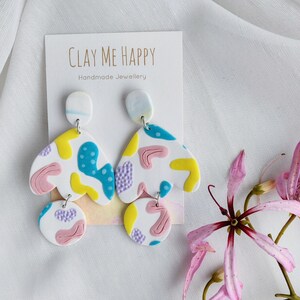 Pode incluir: Um par de brincos abstratos de argila polimérica coloridos. Os brincos são brancos com detalhes em rosa, amarelo, azul e roxo. Os brincos estão em um cartão que diz "Clay Me Happy Handmade Jewellery".