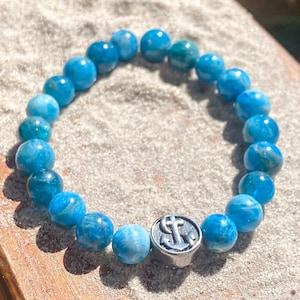 Könnte beinhalten: Ein blaues Perlenarmband mit einem silbernen Anker-Charm. Das Armband hat runde, hellblaue Perlen und einen silbernen Charm mit Anker-Design. Das Armband wird auf einer sandigen Oberfläche präsentiert.
