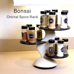 May include: A white and black Bonsai Orbital Spice Rack with three tiered circular shelves holding clear glass spice jars with black lids and circular labels. The text Bonsai Orbital Spice Rack is visible in the upper left corner of the image.